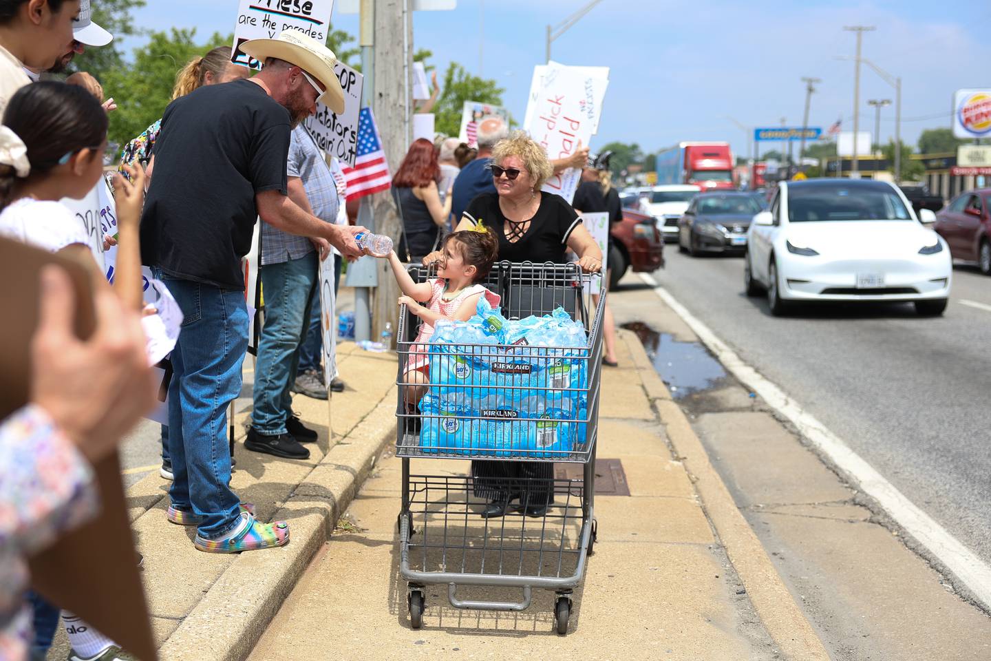 A little girl sits in a cart handing out water to protesters along North Larkin Avenue near West Jefferson Street during the No Kings Day protest on Saturday, June 14, 2025 in Joliet.