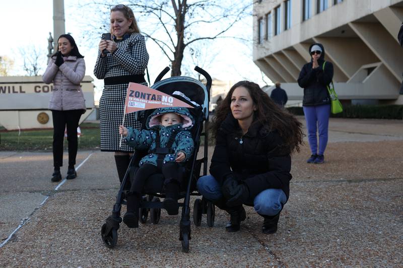 Vivian Foreman, 1 years old, waves a flag as her mother, Amirrah Abou-Youssef, listens to speakers during a rally for ZONTA Says No To Violence Against Women outside the old court house on Tuesday in Joliet.