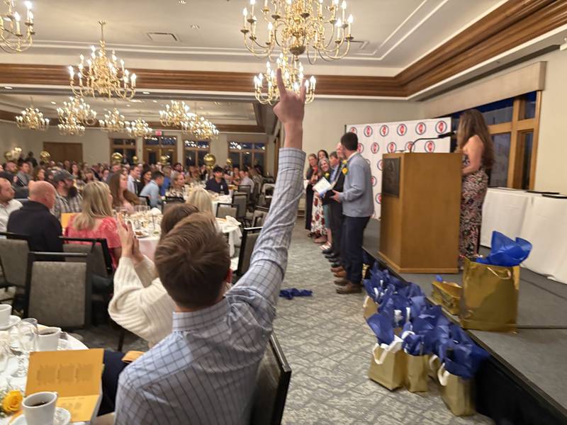 John Aubert holds up a "rock on" sign as Dane Erbach wins the High School Teacher of the Year at the McHenry County Educator of the Year ceremony in Lake in the Hills, Saturday, May 9, 2026.
