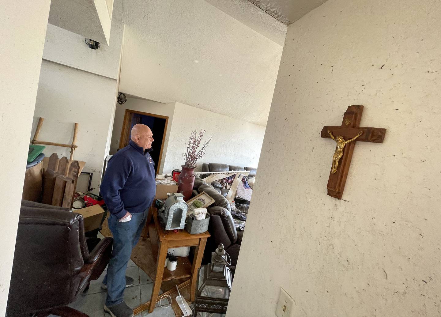 Aroma Township resident Tim Pinson stands among the damage in the living room of his home on Elmwood Drive on March 19, 2026 following the March 10 tornado. Pinson said the few decor items that didn't budge were the crucifixes on their walls.