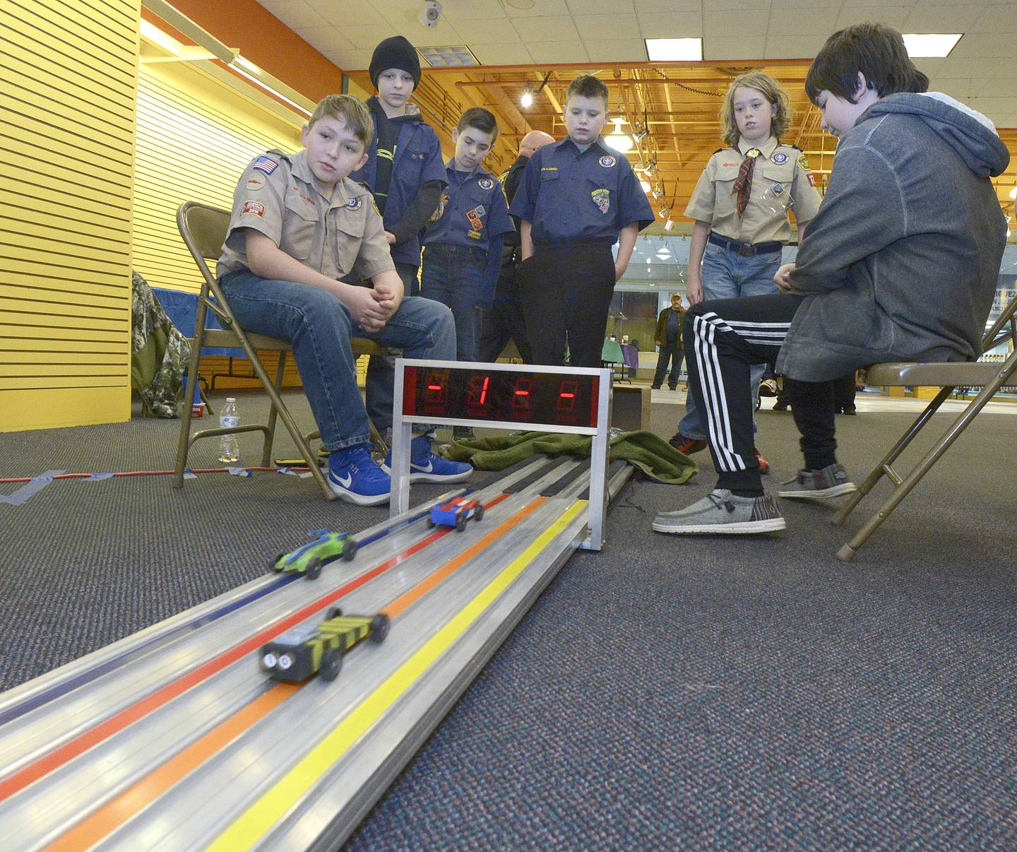 Seated Judges Owen Degroot and Eli Washkowiak watch the pinewood cars cross the finish line, racers, Declan DeRubeis,nicolas Pitstick, Brayden Franklin and Owen Gifford watch to see how their entries finished.