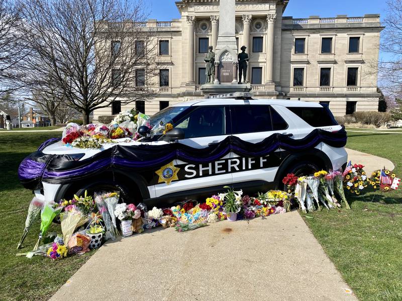 Dozens of flower arrangements, trinkets and other tokens are laid near a DeKalb County Sheriff's Office vehicle on the DeKalb County Courthouse lawn Saturday, March 30, 2024, in memory of sheriff's deputy Christina Musil, who was killed while on duty after her squad car was rear-ended by a truck Thursday, March 28, 2024.