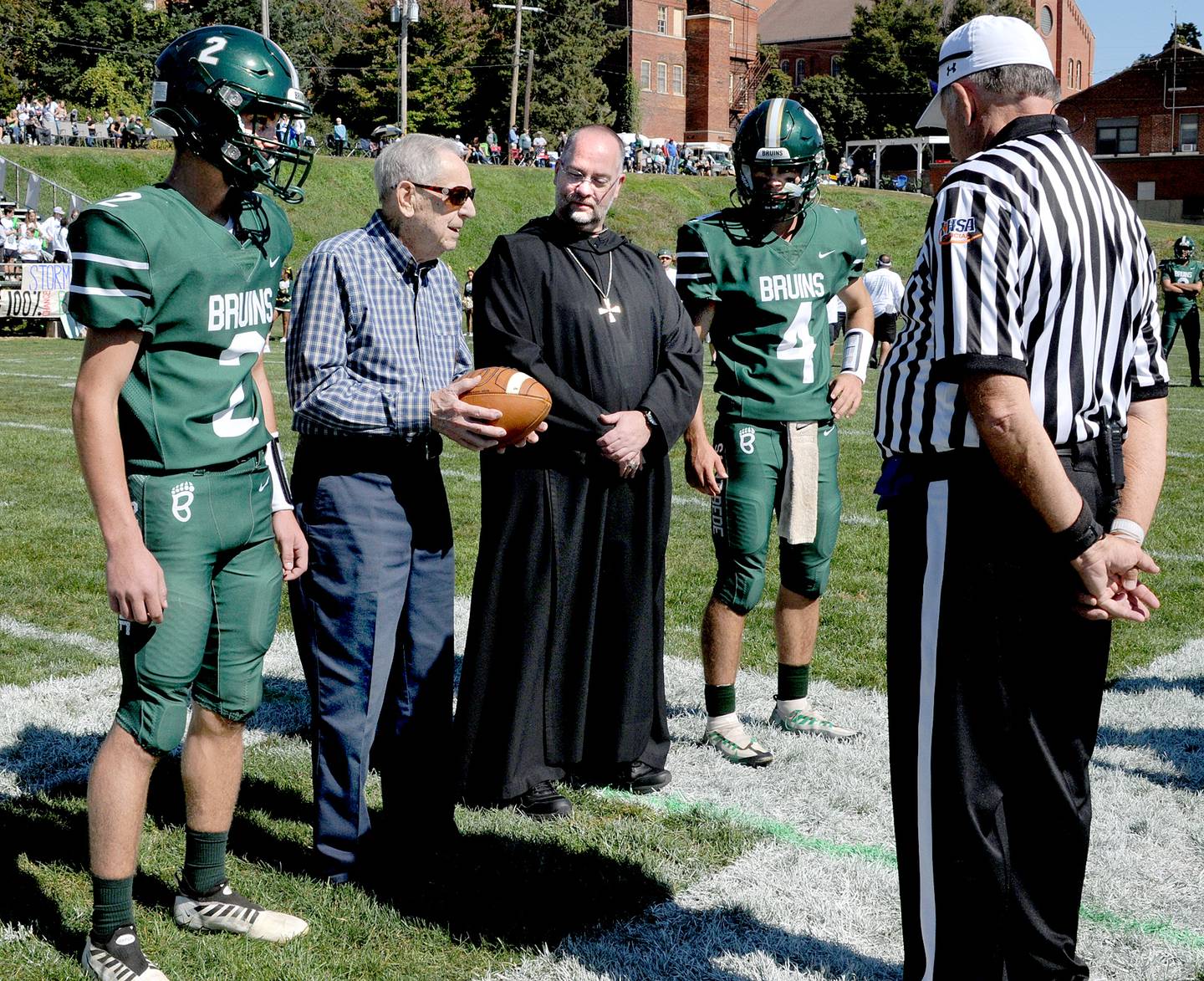 Melvin Baima, who is a 100 year old from the St Bede class of 1939, presents the game ball as his two great grandsons Ryan and John Brady along with Abbot Michael Calhoun look on Saturday, Oct. 1, 2022 before the Homecoming game against Bureau Valley.
