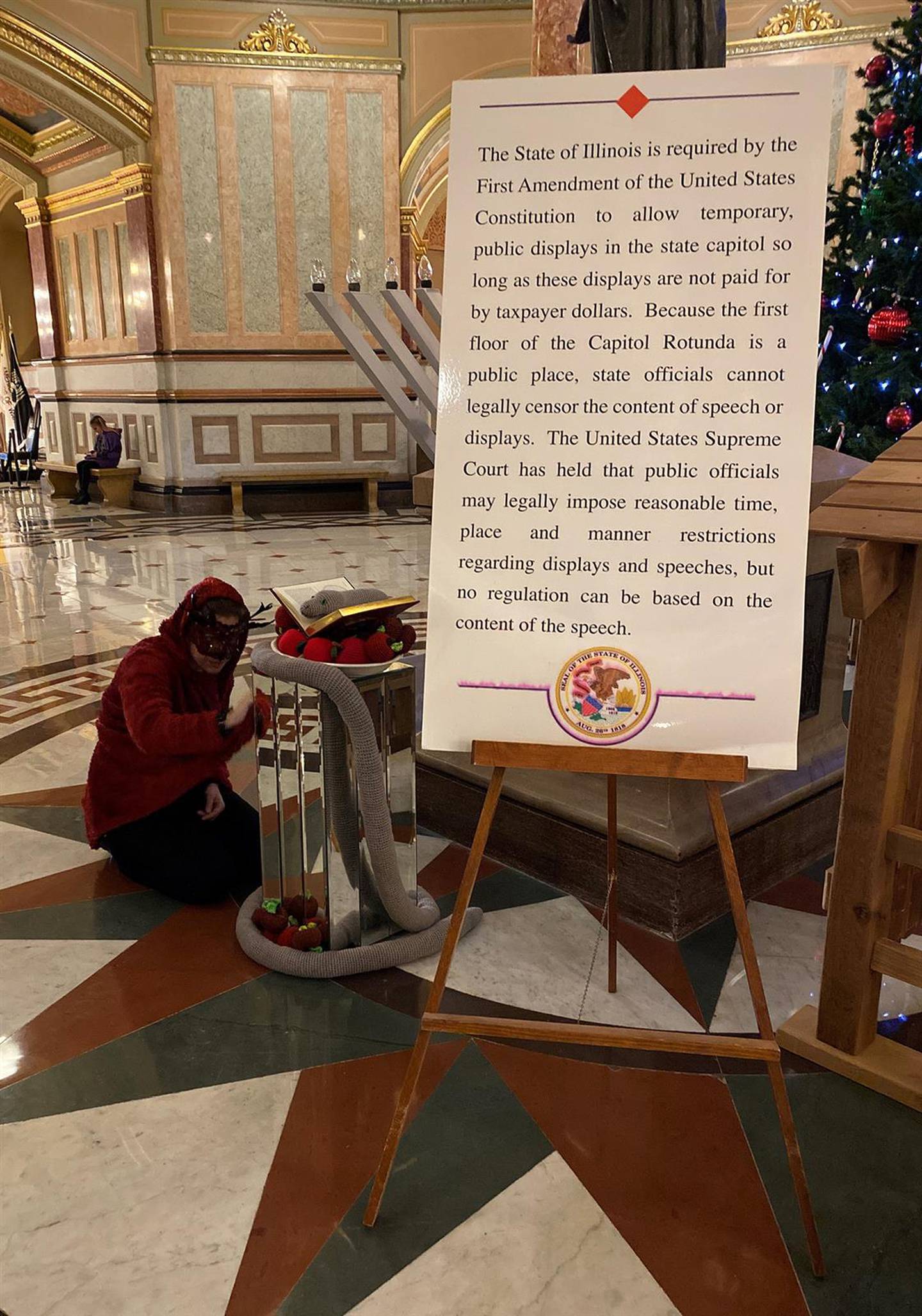 A member of the Satanic Temple of Illinois polishes the group's 2022 holiday display on the Illinois Capitol rotunda. The display consists of a crocheted snake sitting on a book and a pile of apples crocheted by Temple members. (Capitol News Illinois photo by Jerry Nowicki)