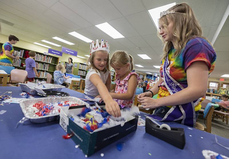Parker Halfacre (left), 8 reaches across sister Natalie, 5, for an adornment as they craft a crown with Petunia Attendant Elyse Stevens Wednesday, July 3, 2024 at the Dixon library. Kids were invited to come in and work on several types of crafts with the Petunia royalty.