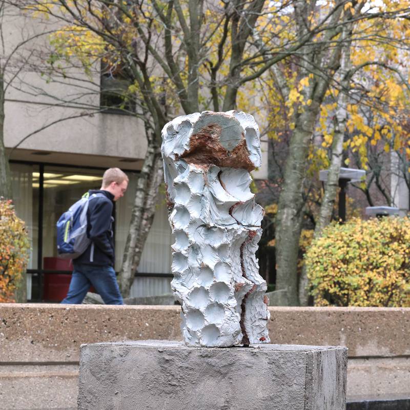 A student passes by a portion of the sculpture "Foundations of Character" by Erinn Stuart Wednesday, Nov. 8, 2023, outside the Northern Illinois University School of Art and Design, Visual Arts Building. The piece is part of the Terrain Biennial 2023, a countrywide exhibit where artists display their work outside of homes and building around town.