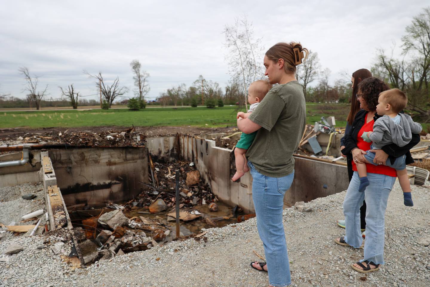 Erin Boerschig, right, and her 8-month-old son Sawyer, stand with Lisa Gerth, holding her grandson Everett, almost 2, next to her daughter-in-law Abigail Gerth, at right, on April 15, 2026, as they look down into the basement where they sheltered during the March 10 tornado that hit Aroma Township.