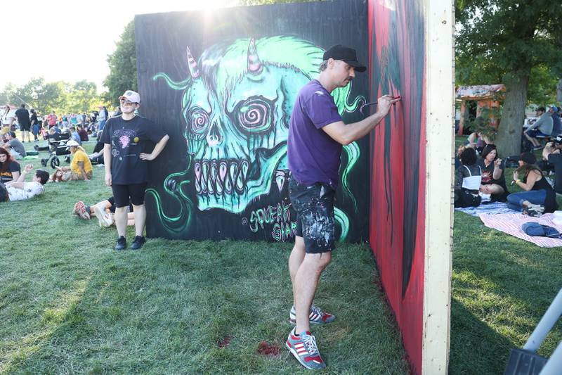Artist Pat Marek works on a painting while a festival goer has her photo taken with his other painting on day one of Riot Fest, Friday, Sept. 15, in Chicago.
