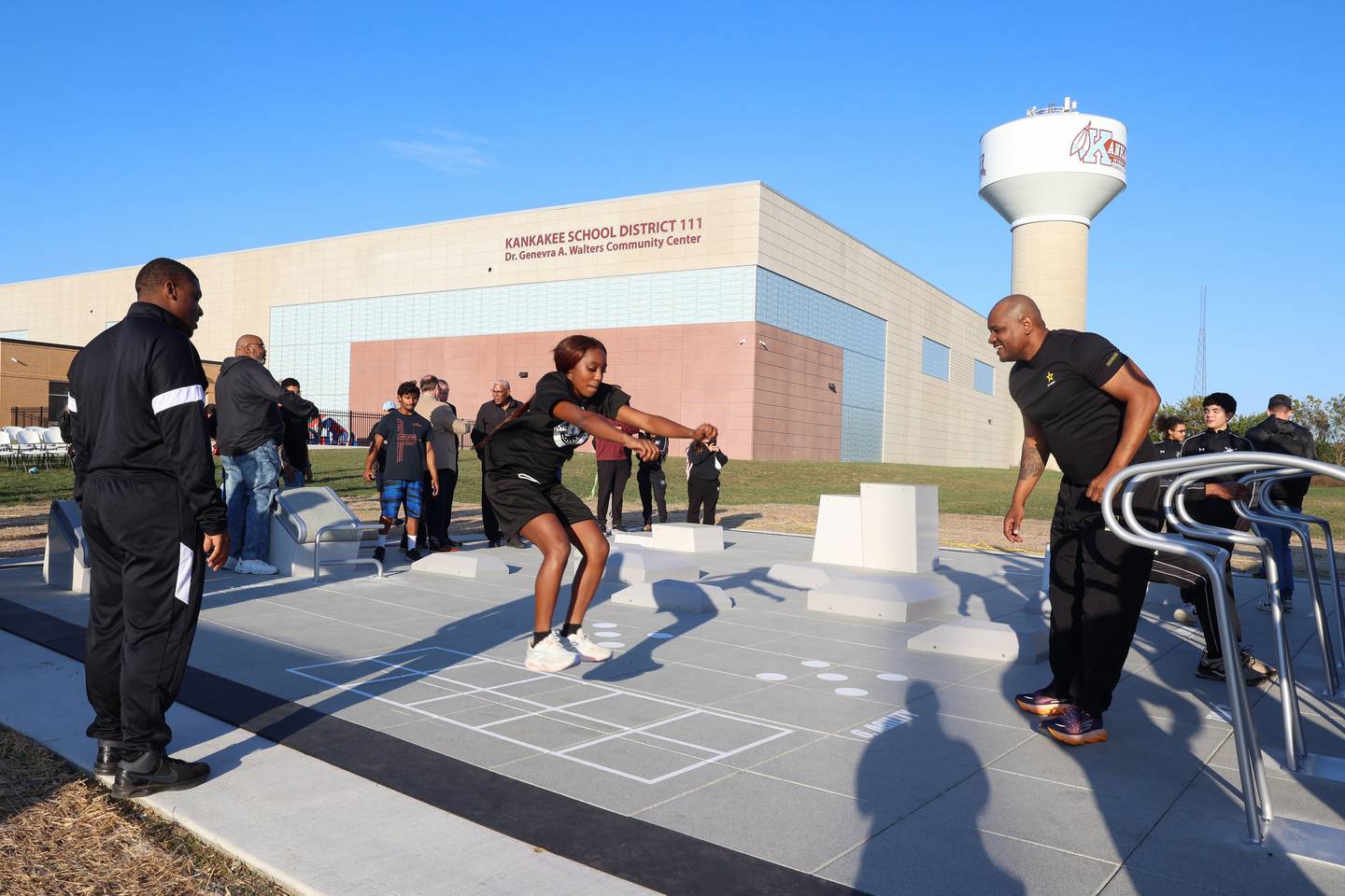 Kankakee's Jasyia Wesby, center, demonstrates an exercise on the new public outdoor fitness court at Kankakee High School as JROTC Army Instructor Marcus Brown, right, cheers her on Thursday, Oct. 16, 2025, after the ribbon cutting ceremony.