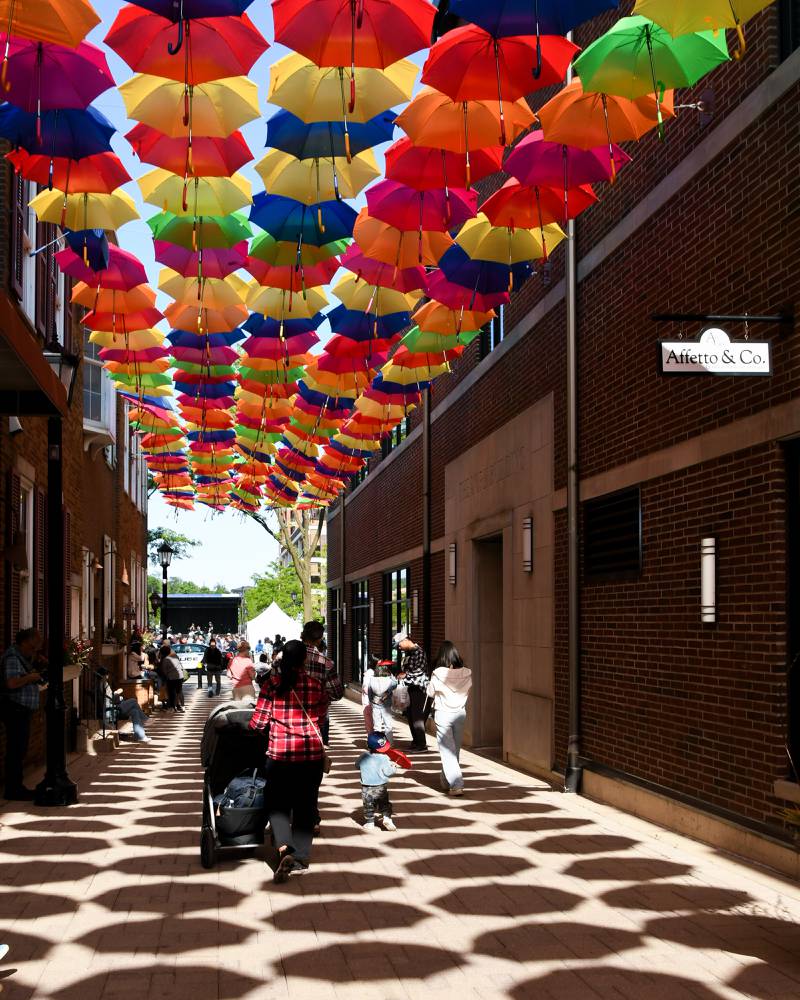 Hanging umbrellas cast a shadow down Schiller Court on Saturday May 31, 2025, as people stop and enjoy taking photos of the umbrellas over head during the Elmhurst Summer Kick Off Party held at City Centre Fountain Plaza in Elmhurst.