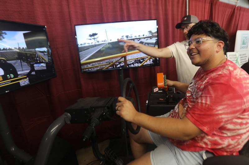 Nickolas Parra, gets directions from Connor Hessman as he drives a farm field sprayer simulator during the opening day of the McHenry County Fair on Tuesday, July 29, 2025. The fair runs through Sunday at the fairgrounds in Woodstock.