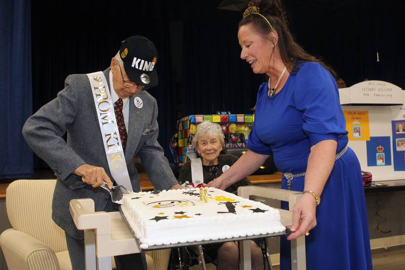 Bob Anderson stands next to his wife, Vivian, and Toni Galster, HOA director, as he cuts the anniversary cake after both he and his wife were crowned Prom King & Queen during the Senior Prom to celebrate the 50th Anniversary of Leisure Village in Fox Lake.