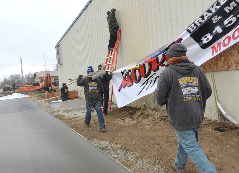 Cement workers from Hendricks Construction walk past Kyle Moore and Todd Billings as they hang a Moore Tires banner on Tuesday, Feb. 6, 2024. The business is moving to a new location after a fire last month destroyed its Rock Falls location.
