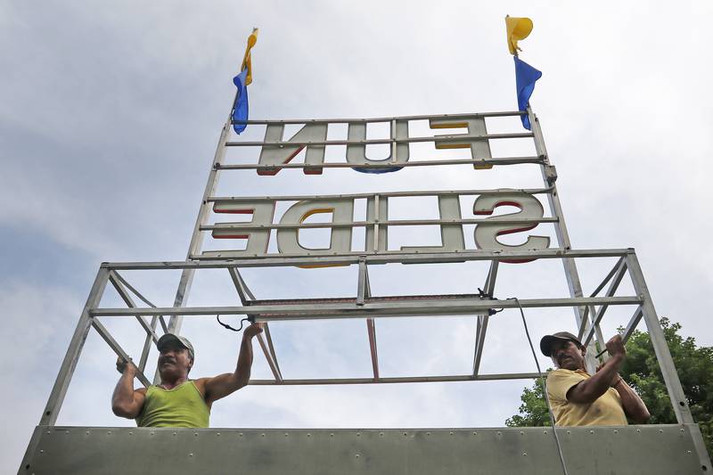 Employees Roque Aurrea, left, and Ramon Mendoza, with Windy City Amusements, hoist up the Fun Slide sign as workers prepare for the opening of this year's Lakeside Festival at the Dole and Lakeside Arts Park on Wednesday, June 30, 2021, in Crystal Lake. Lakeside Festival will run 3 to 11 p.m. Thursday and then noon to 11 p.m. on Friday, Saturday and Sunday.