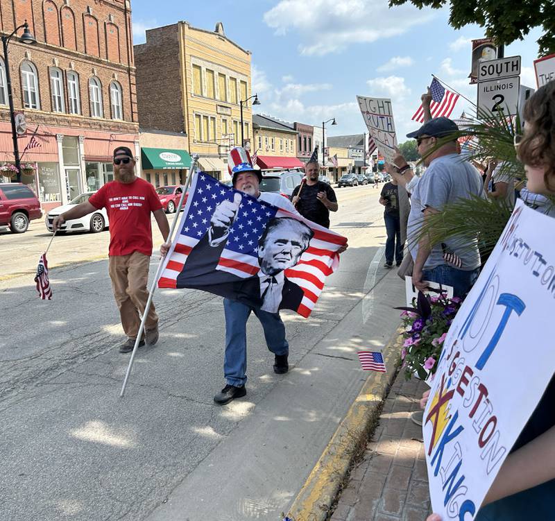 Gary Schrimpser of Oregon, carries a flag in support of Donald Trump as he walks past attendees of the "No Kings" rally on Saturday, June 14, 2025 in Oregon, Illinois. Schrimpser organized a "Flag Day" event in support of Trump while members of Indivisible of Ogle County held their fourth rally protesting Trump's actions. The "No Kings" rally, held on two sidewalks surrounding the historic Ogle County Courthouse, drew an estimated crowd of 500 while the "Flag Day" event had around 10 people attending.