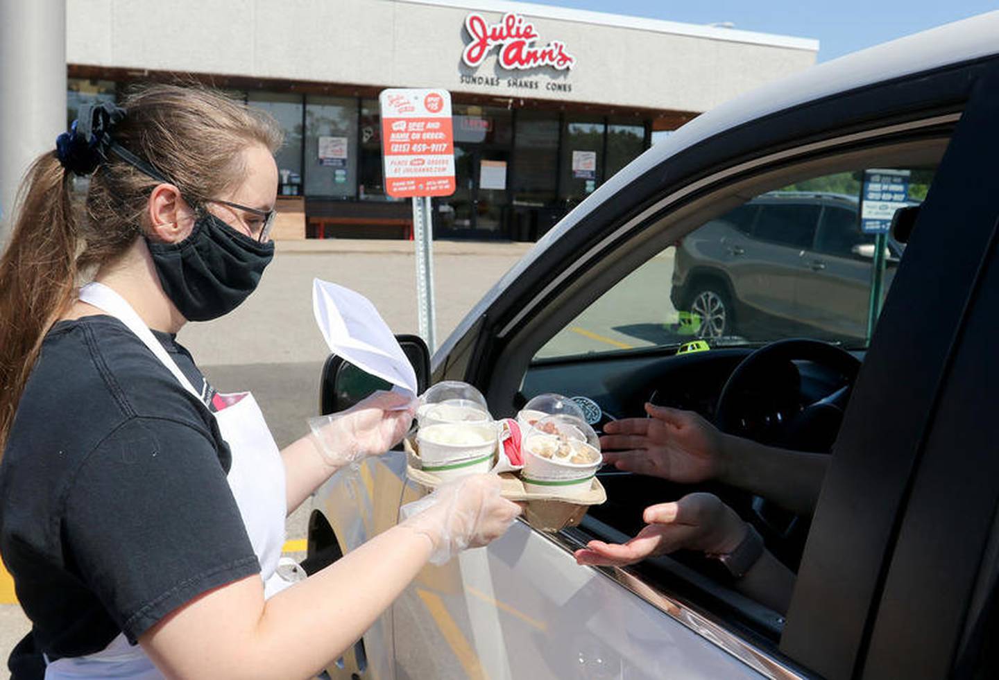 Julie Ann's Frozen Custard carhop and scooper Halle Baldwin of Crystal Lake (left) hands Sarah Pabbathi of Cary her order on Tuesday, June 2, 2020, in Crystal Lake. The National Weather Service listed a high of 96 degrees Tuesday. The business has seen about a 30% increase in sales this year despite social distancing restrictions closing the inside to customers, and has been able to add 6 new employees this year while using a call service through Amazon to handle an increase in phone calls.