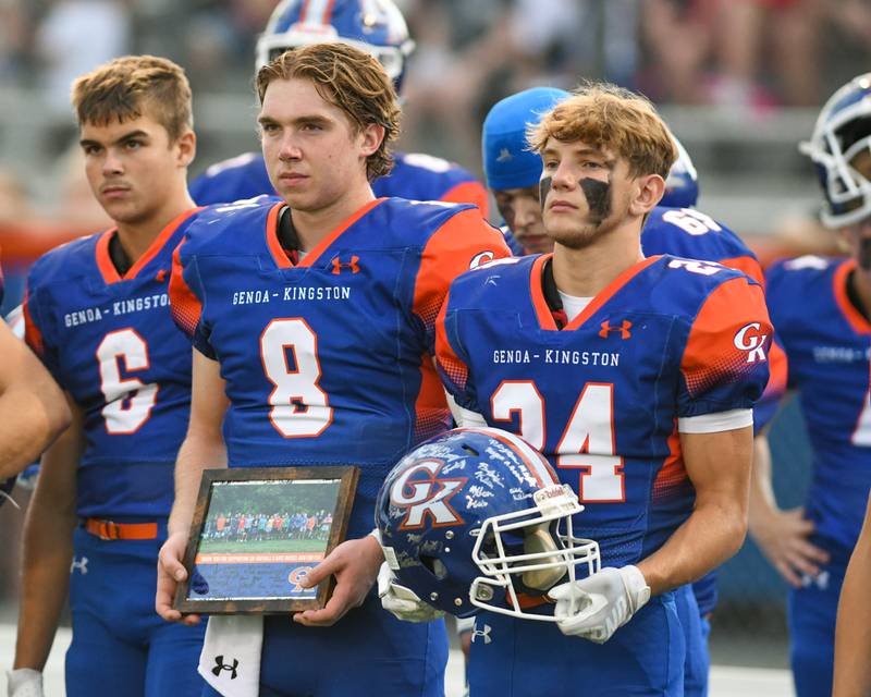 Genoa-Kingston's Patrick Young (24) and teammate Genoa-Kingston's Nathan Kleba (8) bring a sign helmet and photo to the Russell family at the start of the Friday Aug. 30, 2024 game held at Genoa-Kingston to honor former coach David Russell who passed away in December.