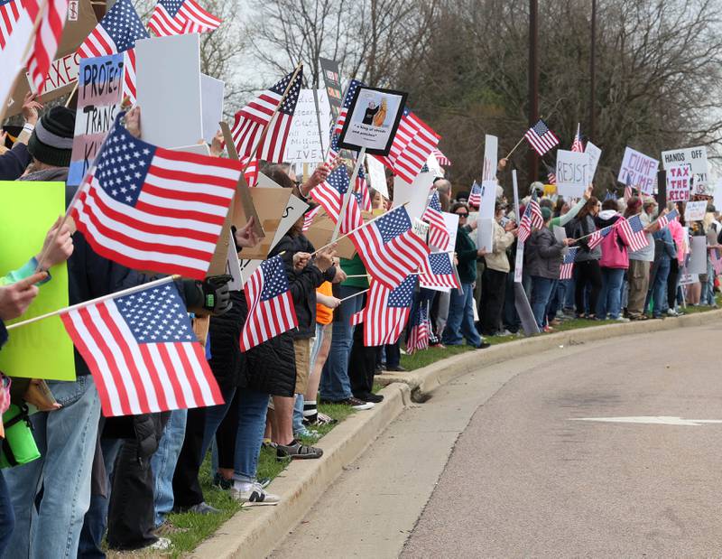 Protesters wave flags and hold signs along Sycamore Road in front of Hopkins Park Saturday, April 19, 2025, during a “No Kings” rally and march in DeKalb. The group was protesting what they’ve argued is the unlawful and harmful federal policies of President Donald Trump and his administration.