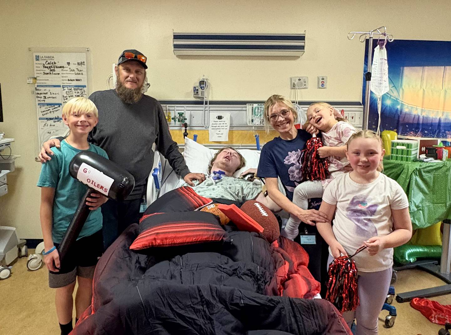 Jennifer and Nick Outerbridge and their three youngest children, Noah, 11, Ella, 8, and Gracie, 5, pose for a photo with their oldest son Caleb, now 17, in his hospital room, decorated to celebrate his football senior night in October. Caleb, who suffered a rare autoimmune disease just before last Thanksgiving that rendered him unconscious, has made progress showing signs of awareness and interaction.