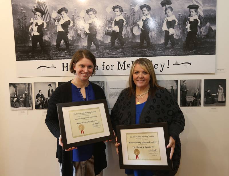 The Bureau County Historical Society has received statewide recognition for its work preserving and sharing local history. Jessica Grey curator and Research Associate Erica Oertel marketing coordinator pose for a photo with the awards on Friday, May 1, 2026 at the Bureau County Historical Society in Princeton. 
The Illinois State Historical Society recently named the organization’s publication, The Chronicle, and its Henry W. Immke Exhibit as 2026 “Best of Illinois History” award recipients.
The Henry W. Immke Exhibit highlights the work of Henry W. Immke, a longtime Princeton photographer who operated a studio in the community from 1866 to 1923. Immke is believed to have taken more than 30,000 photographs during his career, documenting life in Bureau County and the surrounding region.
Over the course of more than four decades, the museum digitized Immke’s extensive collection. Today, more than 22,000 of those images — including glass plate negatives, daguerreotypes and tintypes — are preserved at the History Center. The Immke exhibit is open to the public Friday and Saturday afternoons through July at the Newell-Bryant Museum at 634 Pleasant Street in Princeton. In addition to the exhibit, The Chronicle continues to expand its reach. The publication now is distributed to readers in 49 states and 89 Illinois cities.