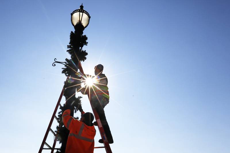 Jake Hanetho, top, and Robert Altman with the Public Works Department set up garland and lights around light posts lining N. Main St. on Thursday, Nov. 4, 2021 in downtown Crystal Lake.