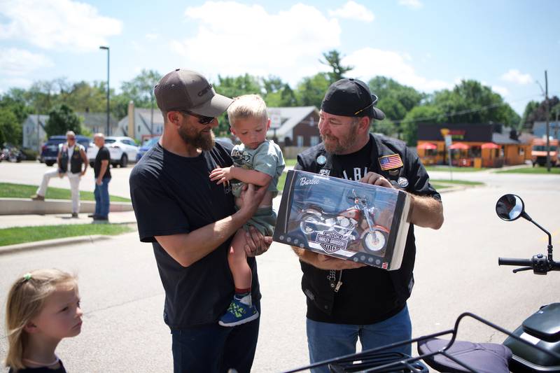 Oilver Olinger, 2, held by Dad Nathan Olinger, receives a gift from Donnie Williams of American Legion Planfield Post No. 13 outside Sycamore Public Library on Sunday, June 22, 2025. Oliver was escorted by law enforcement and bikers to the St. Charles Motorcycle Museum to celebrate his cancer remission.