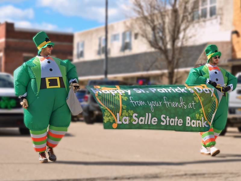 St. Patrick's parade participants wearing inflatable leprechaun suits hold a banner on Saturday, March 9, 2024, on First Street in La Salle.