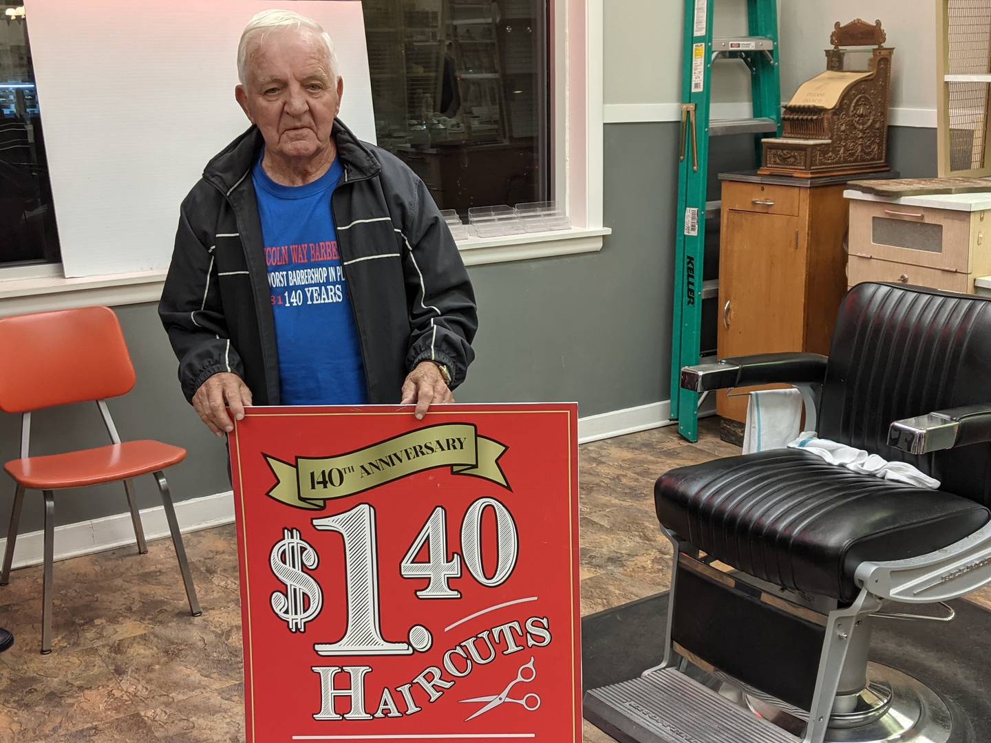 Lincolnway Barbershop owner Don Kinley holds a sign from when the shop celebrated its 140th anniversary in 2021 and offered $1.40 haircuts to customers.