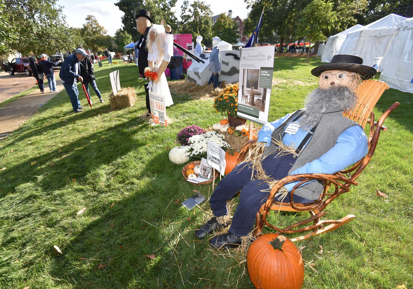 Visitors look closer at a bride and groom scarecrow as another, sponsored by Amish Firniture store sits in a wooden rocking chair at the Annual Scarecrow Festival in Lincoln Park in St. Charles on Friday, October 6, 2023. The event runs through the weekend with family activities and a chance to vote for your favorite scarecrow.