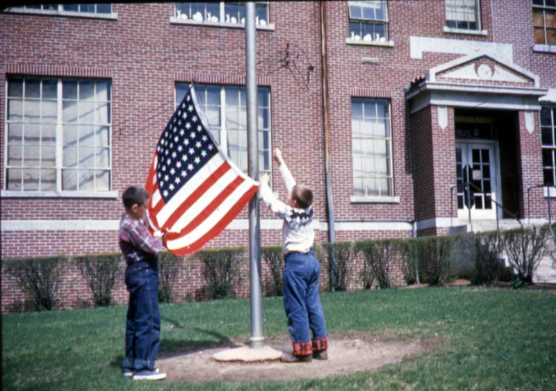 Oswego fifth graders Mike Ode (left) and Bob Chada raise Old Glory at the old Red Brick School in the spring of 1957. The site of the building at Madison and Jackson streets, which opened in 1886 and was demolished in 1965, will be one of those participants will visit during the Oswegoland Heritage Association’s “Oswego History Walk—Schools” this Sunday, Aug. 28.