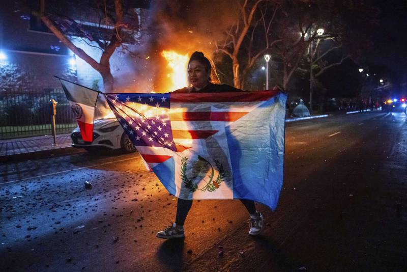 A person displaying multiple flags walks past a burning car during protests over the Trump administration's immigration raids in Los Angeles, Monday, June 9, 2025. (AP Photo/Ethan Swope)