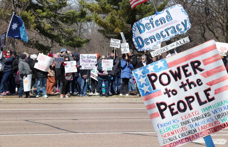 Hundreds of protesters hold signs and chant slogans along Sycamore Road in front of Hopkins Park in DeKalb Saturday, March 28, 2026, during a No Kings march and rally against the federal actions of President Donald Trump and his administration.