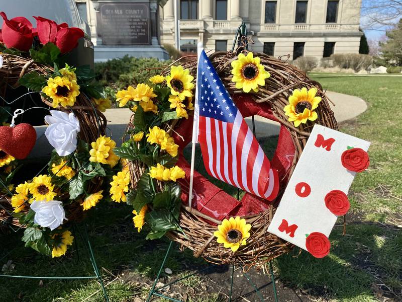 A floral wreath with the word "Mom" on it stands next to a DeKalb County Sheriff's Office vehicle displayed on the DeKalb County Courthouse lawn in Sycamore on Saturday, March 30, 2024. Mourners have visited the site to leave trinkets, flowers and other tokens in memory of sheriff's deputy Christina Musil, who was killed while on duty after her squad car was rear-ended by a truck Thursday, March 28, 2024.