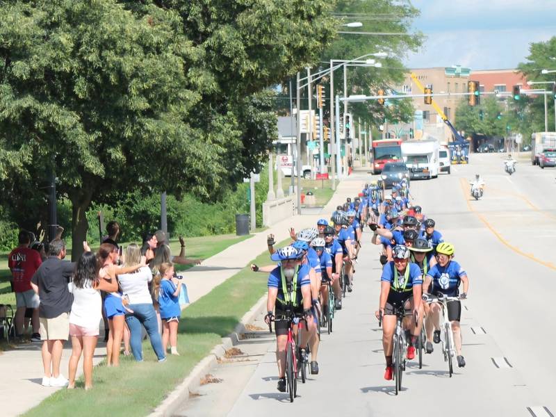 Onlookers wave to bicyclists as they make their way through downtown DeKalb along Lincoln Highway (Illinois Route 38) on Saturday, July 12, 2025. Nearly 100 cyclists biked from Normal to DeKalb on Saturday, July 12, 2025, for Cycle Across Illinois, a charity campaign to honor fallen police officers and raise funds for their families.
