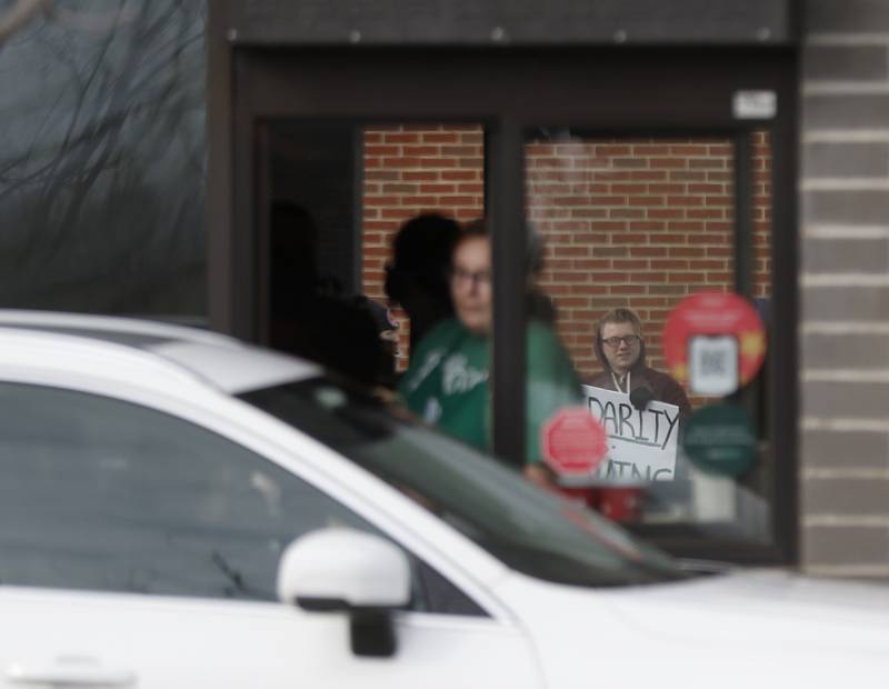 A union picketer is seen through the store as a Starbucks worker hands a customer their order Thursday, Nov. 17, 2022, at the Cary Starbucks location on Starbucks' Red Cup Day. Workers at Starbucks in Cary joined other workers across the country seeking to convince the company to better negotiate with its unionized locations.
