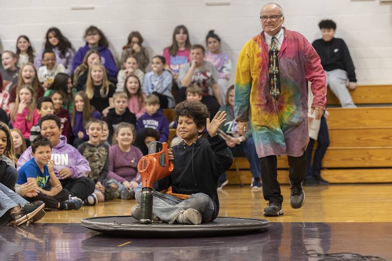 Malachi Pesek waves as he is pushed across the floor on a layer of air by science entertainer Steve Belliveau Friday, March 7, 2025, at Madison School in Dixon. Belliveau was teaching the students about friction.