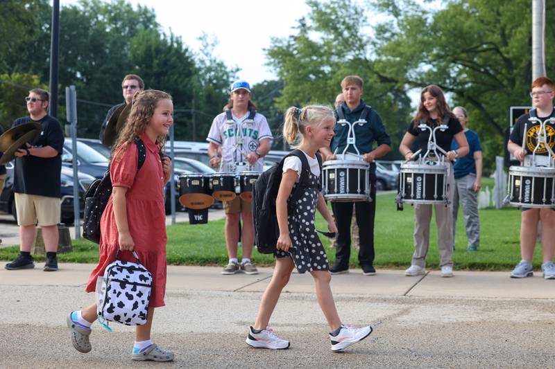 Herscher Intermediate School students Teagan Down, right, and Kaelynne Coulthard smile as they walk past the Herscher High School drumline upon arriving for their first day of third grade on Tuesday, Aug. 19, 2025.