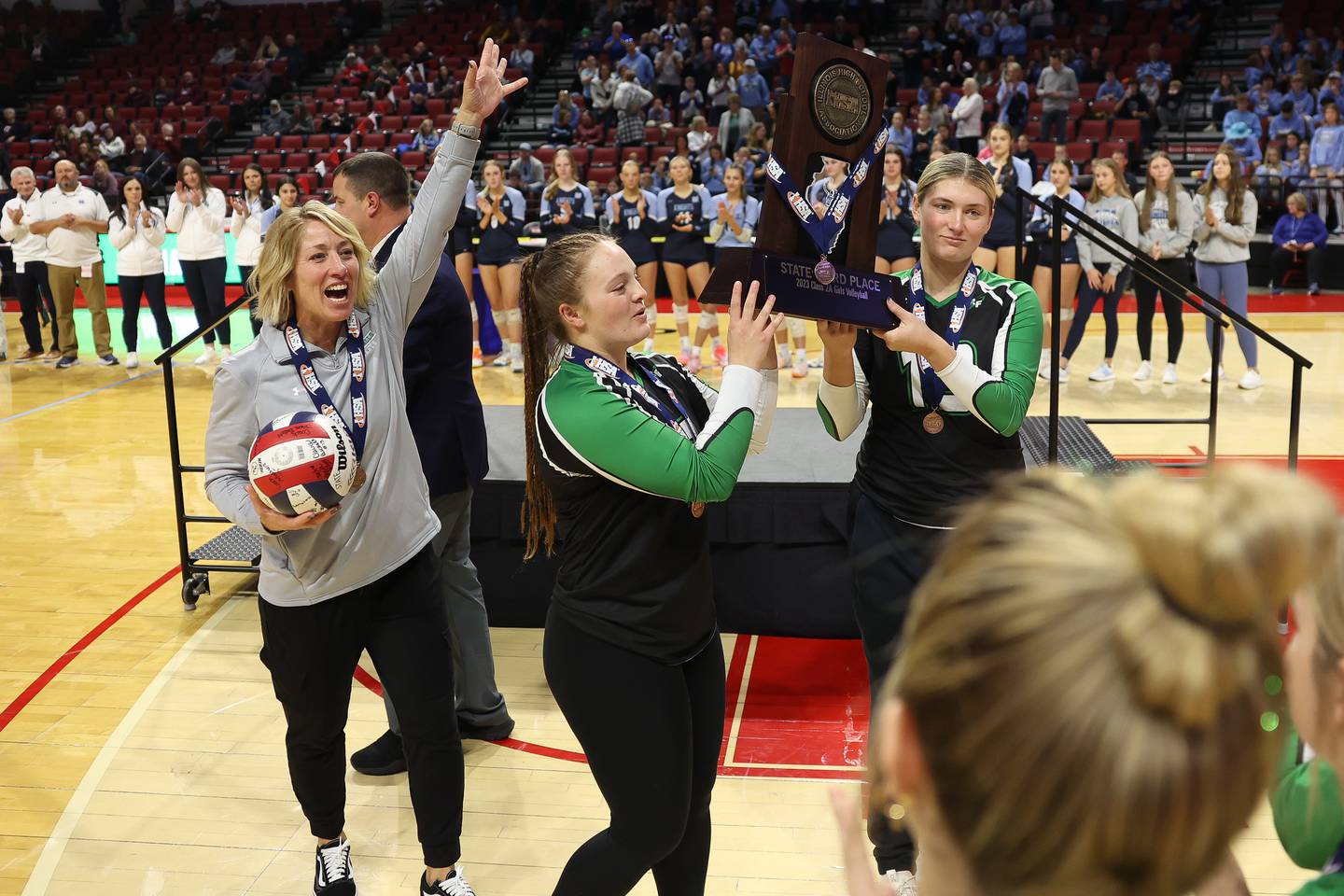 Rock Falls’ team captains Denali Stonitsch, center, and Claire Bickett receive the Class 2A Third Place trophy as head coach Shelia Pillars celebrates after the Rockets win against Carmi-White County in the Class 2A Volleyball Third Place match on Saturday, Nov. 11, 2023 in Normal.