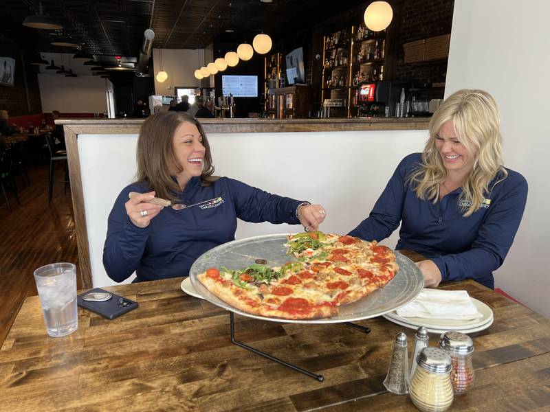 DeKalb County Convention and Visitors Bureau Executive Director Cortney Strohacker (left) laughs as she and part time bureau worker Katherine McLaughlin serve themselves pizza slices at Pizza Beer Whiskey on March 17, 2023.