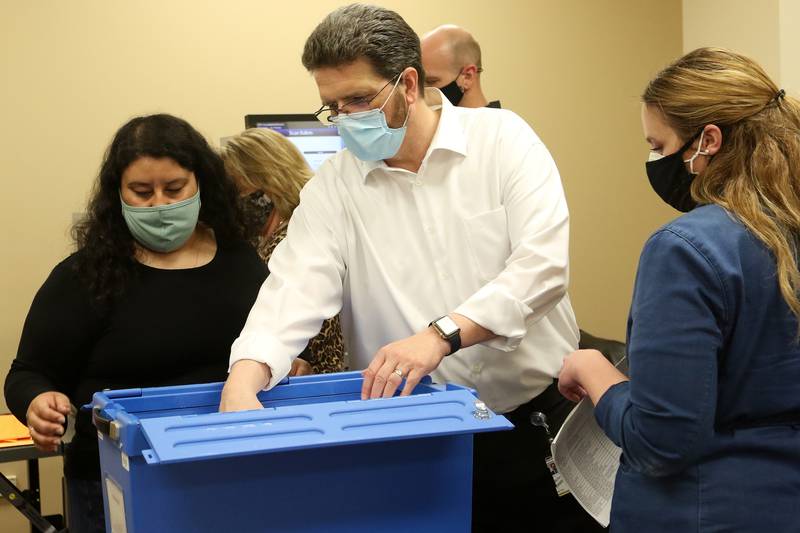 McHenry County Clerk Joe Tirio, center, reaches into a secure blue box for a handful of ballots inside the voting tabulation room at the McHenry County Administrative Building on Thursday, April 8, 2021, in Woodstock.