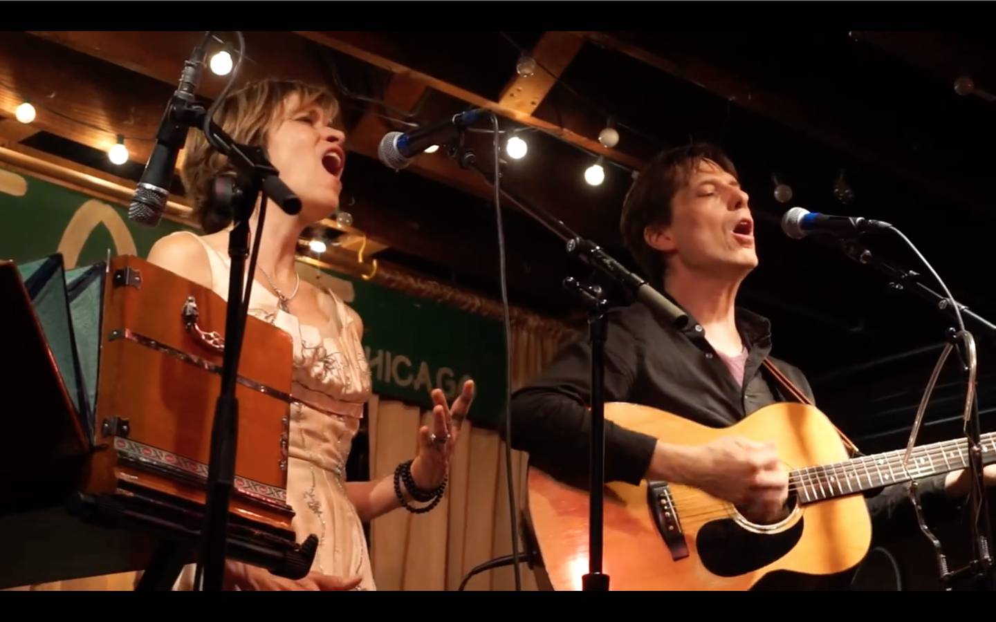 Musicians Diane Christiansen and Steve Dawson standing and playing and singing at a live show. They will be guest artists with guitarist Muriel Anderson, seated with her harp guitar, pauses and smiles between songs at a live show. Anderson will appear Nov. 30 2024 in Downers Grove.