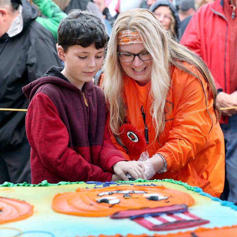 Harrison Rowland, winner of the theme contest, gets some help cutting the first piece of cake from Brandi Voigt, theme director of the Pumpkin Fest Committee, Wednesday, Oct. 25, 2023, on North Maple Street adjacent to the DeKalb County Courthouse during the Sycamore Pumpkin Festival. The theme for this year's festival is Pumpkins of History.