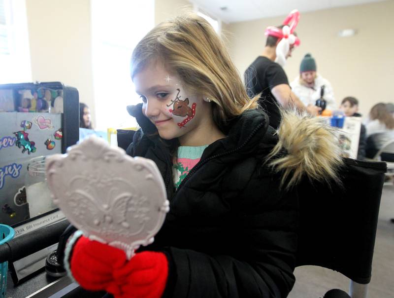Paisley Baryla, 5, of Wauconda looks in a mirror to see the reindeer that was painted on her face in the Community Room at the Wauconda Area Chamber of Commerce during Holiday Walk on Main in Wauconda. The face painting was done by Irisaflower Face Designs.