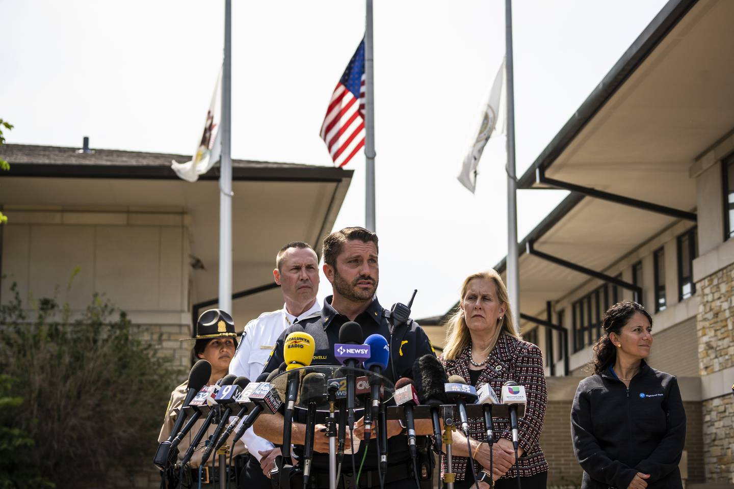 Flag hang at half staff as Highland Park Police Chief Louis Jogmen and Mayor Nancy Rotering look on while Deputy Chief Christopher Covelli, of the Lake County sheriff's office and the Lake County major crimes task force, speaks to the media during a news conference outside the Highland Park Police Department, Tuesday morning, July 5, 2022, in Highland Park, Ill., one day after a gunman killed several people and wounded dozens more by firing a high-powered rifle from a rooftop onto a crowd attending a Fourth of July parade, (Ashlee Rezin/Chicago Sun-Times via AP)