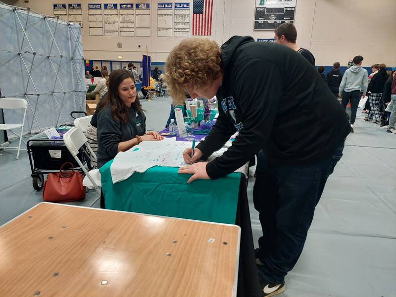 Jeremy Smith Jr., a senior at Princeton High School, signs a shirt that says "I believe you" in support of sexual and domestic violence victims at the Freedom House table Friday, April 19, 2024, during the Career, Job and Volunteer Fair at Princeton High School. Tonya Fitzpatrick (seated) at Freedom House spoke about April being Domestic Violence Awareness Month.