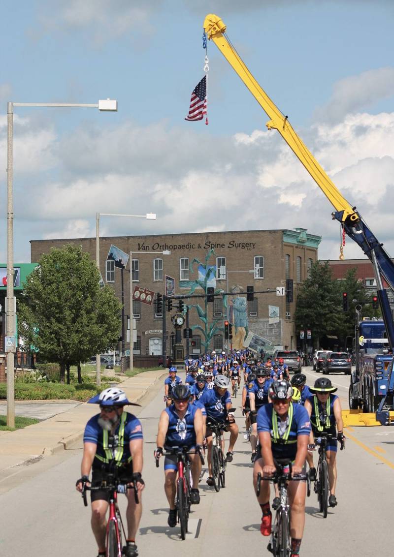 Bicyclists are greeted by an American flag as they make their way through downtown DeKalb along Lincoln Highway (Illinois Route 38) on Saturday, July 12, 2025. Nearly 100 cyclists biked from Normal to DeKalb for Cycle Across Illinois, a charity campaign to honor fallen police officers and raise funds for their families.