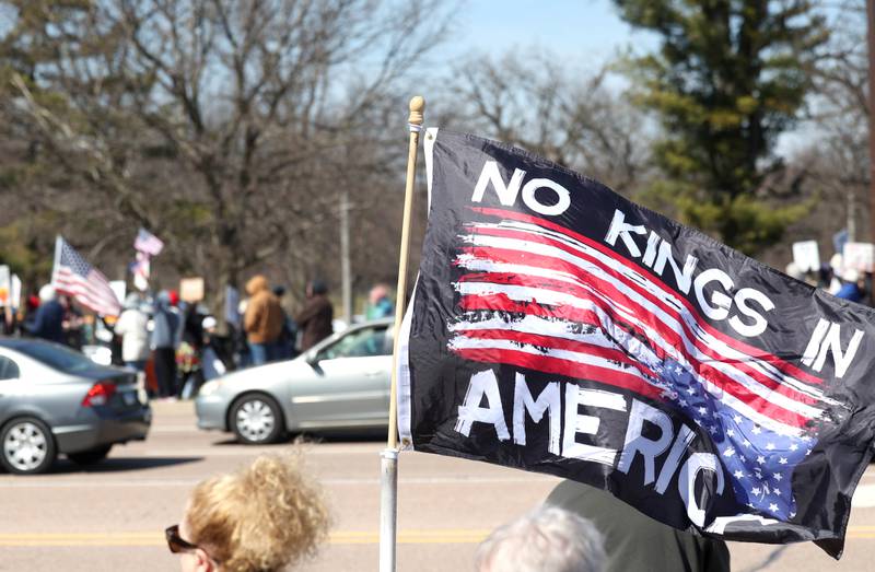 A flag waves in the wind as protesters and vehicles line Sycamore Road in DeKalb Saturday, March 28, 2026, during a No Kings march and rally against the federal actions of President Donald Trump and his administration.