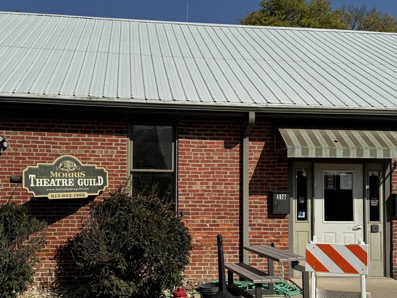 The Morris Theatre Guild in Morris, Ill. It's a brick building with a green tin roof and a sign that reads "Morris Theatre Guild". There are two bushes in front of it.