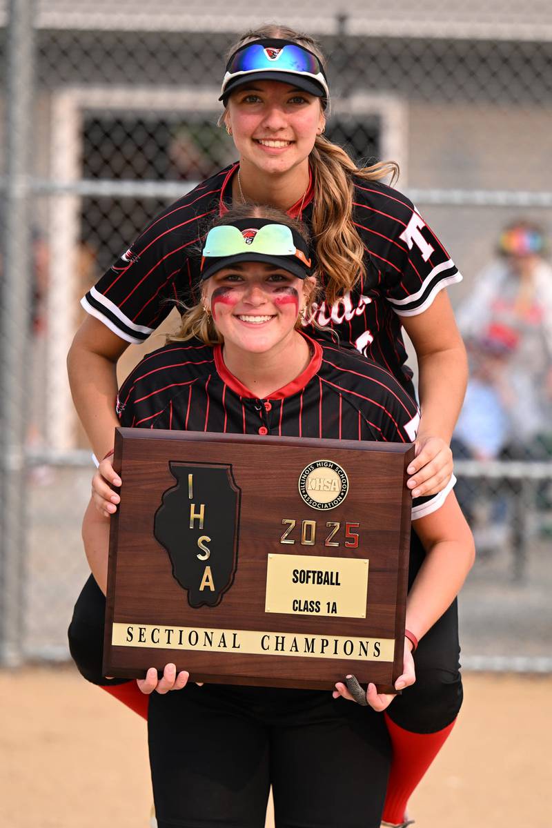 Brooklynn Thompson holds the plaque her Henry-Senachwine High School softball team earned for being sectional champions in 2025, while teammate Rachel Eckert jumps on her back for a picture.