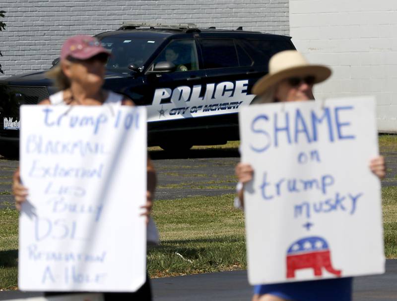 A McHenry police officer watches as Diane Messman and Pattie Klein, both of Woodstock,  protest along Route 31 near of the McHenry Public Library on Friday, June 20, 2025, during a Save Our Libraries Protest hosted by Indivisible McHenry County to draw attention to cuts to the nation’s libraries by the Trump administration.